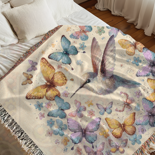 A woven blanket with a floral design featuring hummingbirds and butterflies in pastel colors on a cream background, displayed on a bed.