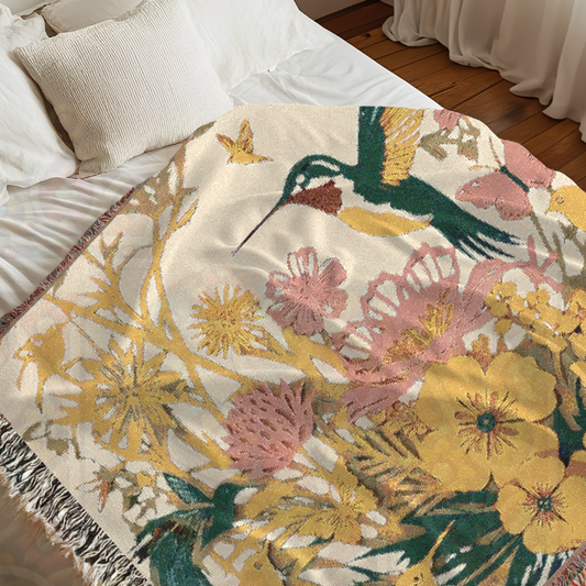 A woven blanket with a floral and bird design draped over a bed, featuring vibrant colors and fringed edges.
